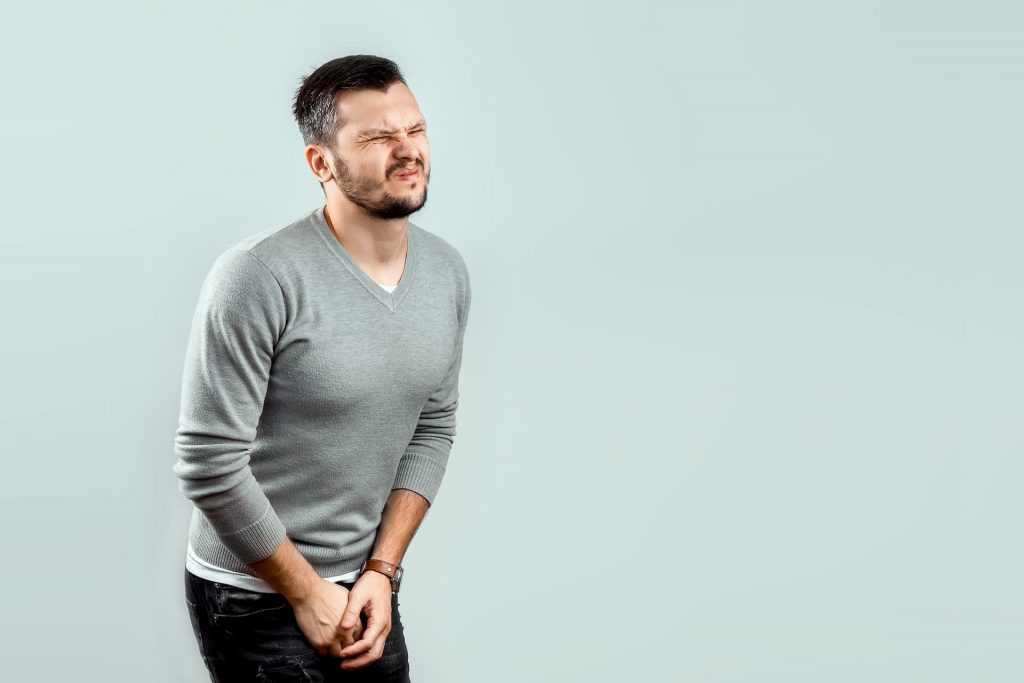 Middle aged man wearing a grey v-neck sweater and black jeans holding his crotch and wincing in pain | Priapism: The Raging Erection You DON’T Want! | e-Surgery