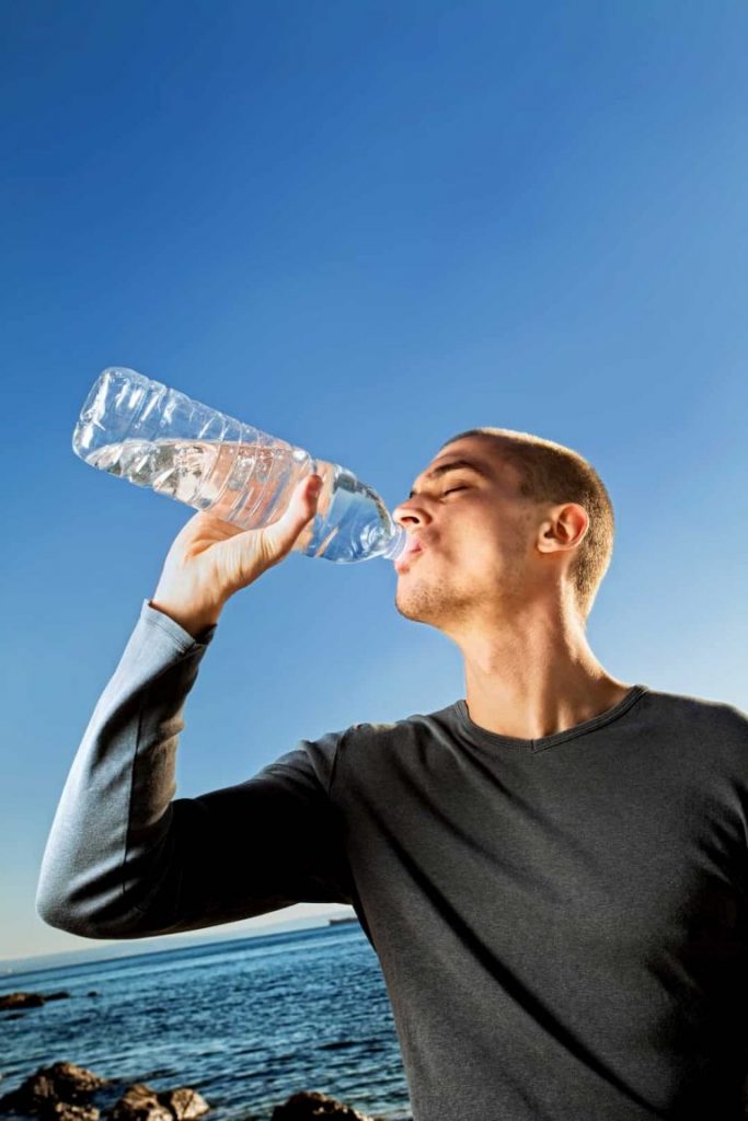 Middle aged bald man wearing a long grey sleeve t-shirt at the beach drinking from a litre bottle of water | Priapism: The Raging Erection You DON’T Want! | e-Surgery