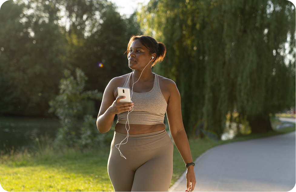 Confident woman in activewear walking through a park while listening to music on her earphones and tracking her progress on a smartphone, embodying the fitness journey and healthy lifestyle transformation